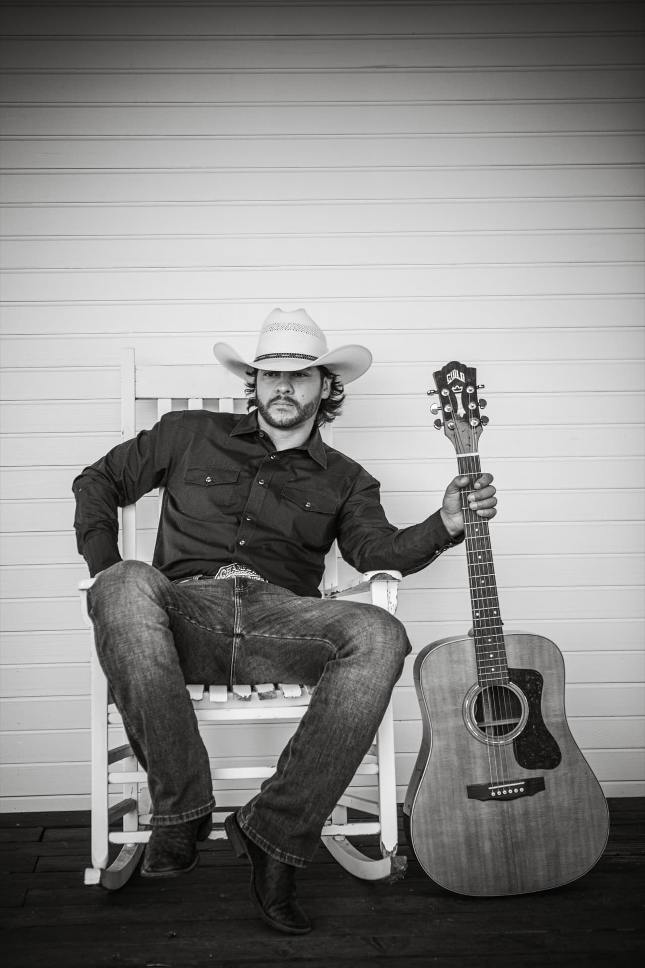 Buck Ford seated in a white rocking chair on a porch, straw hat on, Guild guitar standing beside him