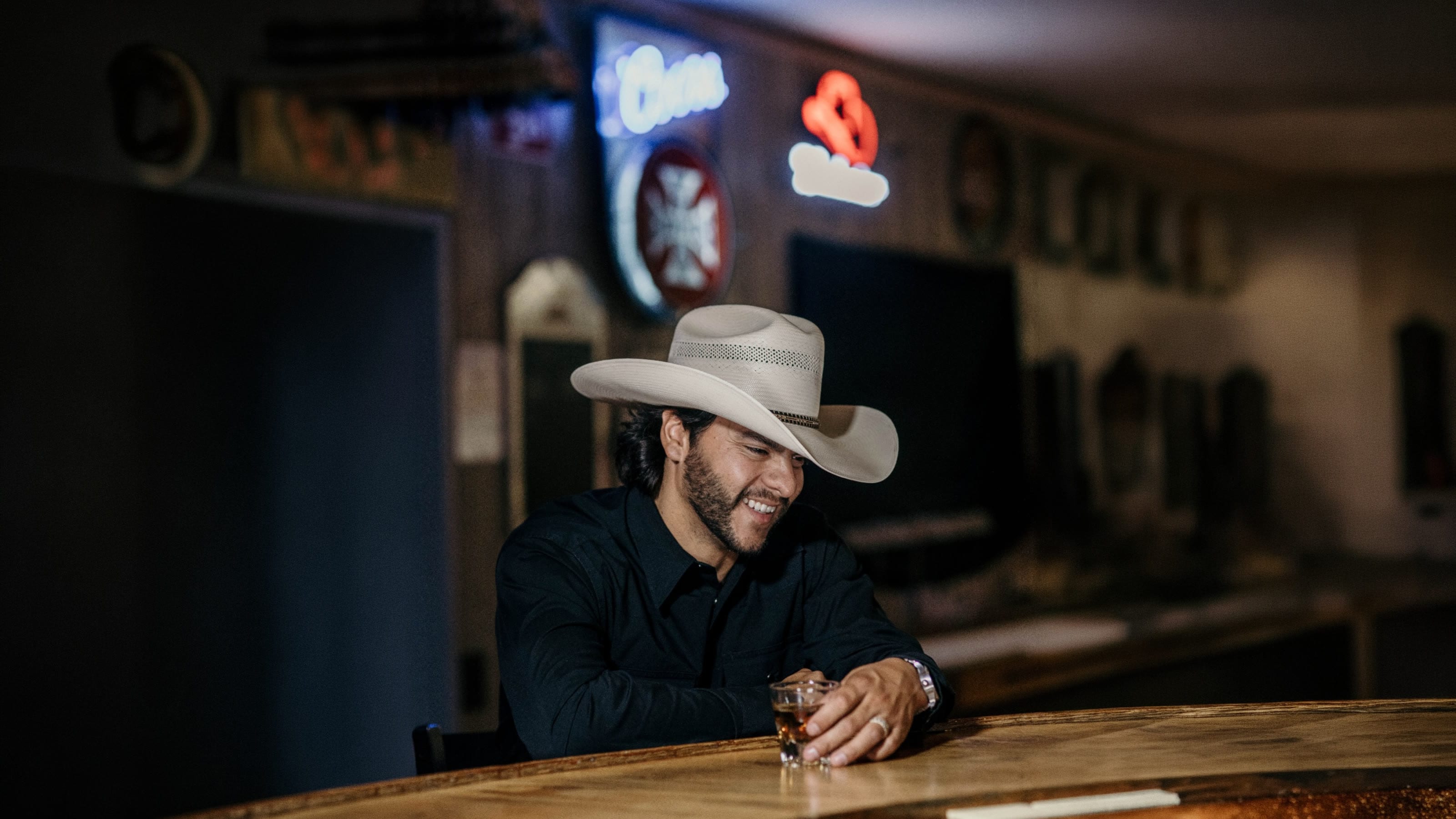 Buck Ford at a honky-tonk bar in a straw hat, whiskey in hand, neon signs glowing behind him